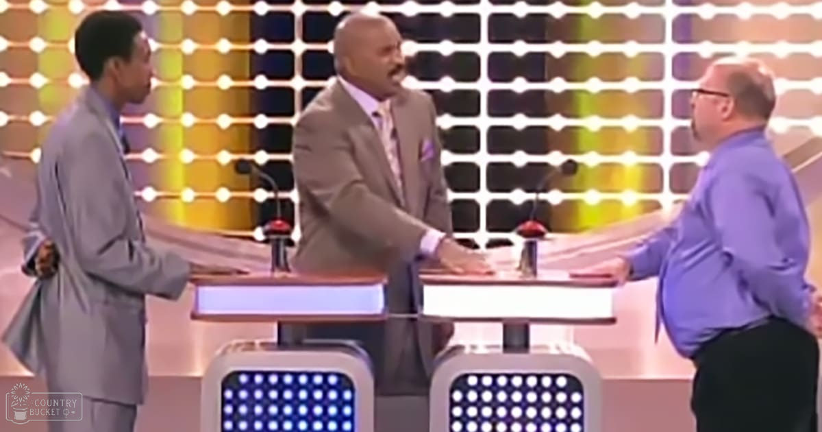 Three adults dressed formally on a game show set with microphones and red buttons, patterned backdrop, beige suits, light gray pants, purple shirt, glasses, well-lit indoor studio environment.