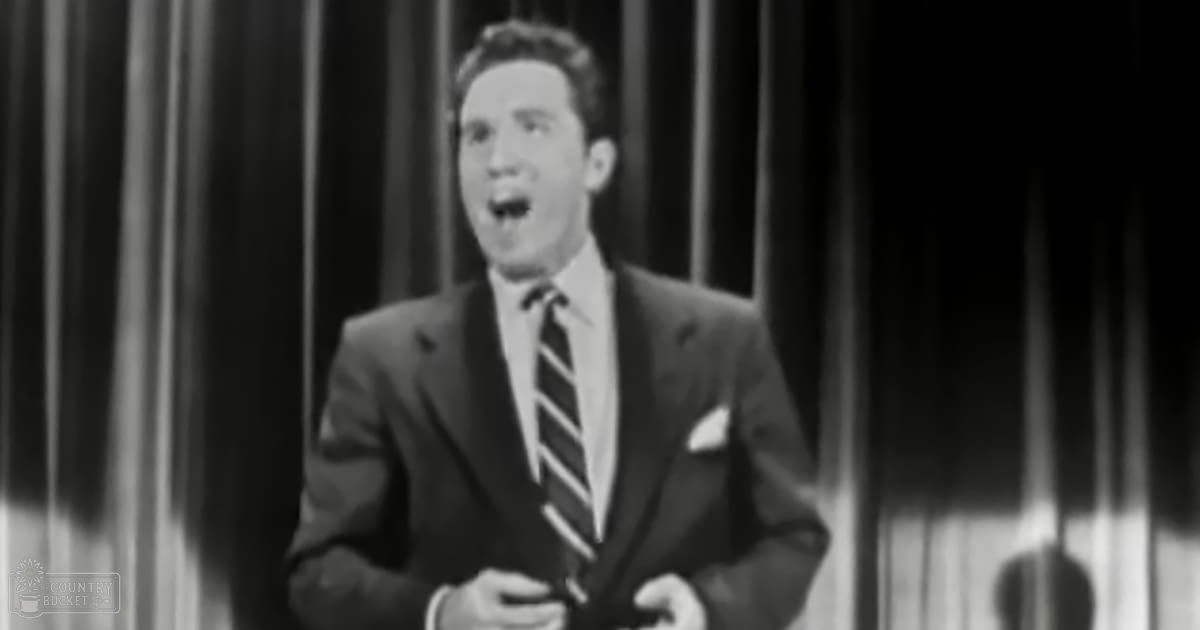 Young adult male wearing a dark suit jacket with diagonal striped tie, mid-20th century style, on an indoor stage at night.