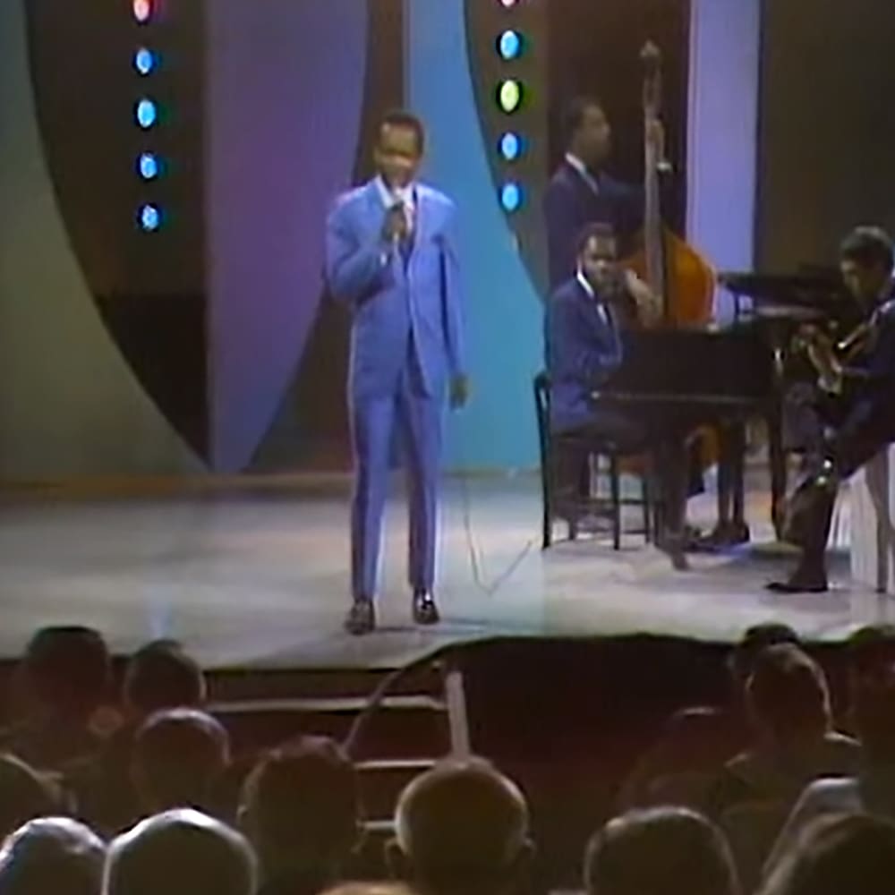 A mid-20th century-style concert scene with a performer in a light blue suit, a pianist in a dark suit jacket, and a saxophonist next to the piano under stage lights.