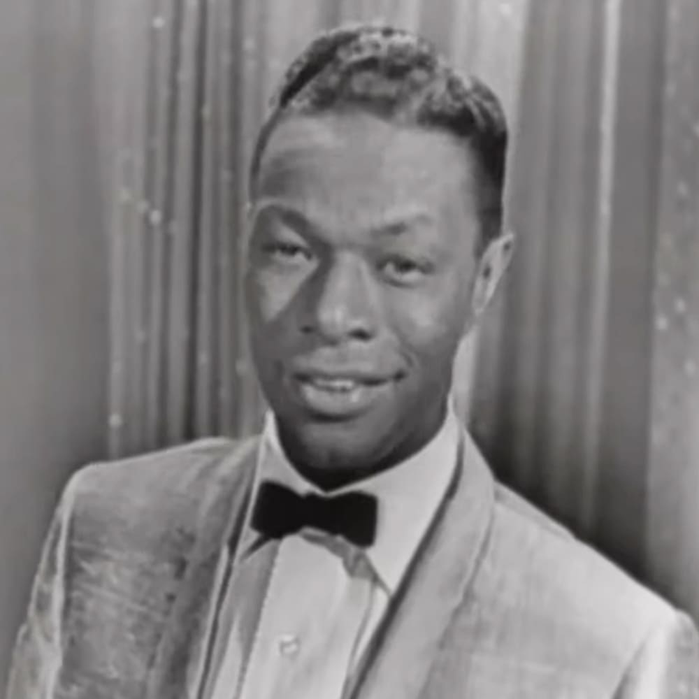 A young adult male wearing a light-colored suit jacket with a bow tie and white collared shirt, set against an indoor backdrop resembling theater drapes.