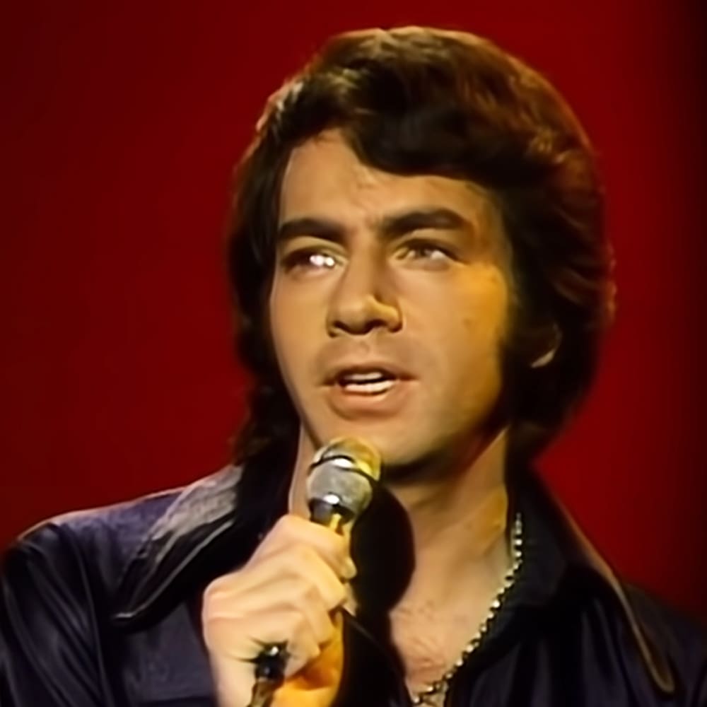 Young adult male singer performing on stage in a dark-colored shirt, holding a vintage-style microphone against a solid red backdrop.