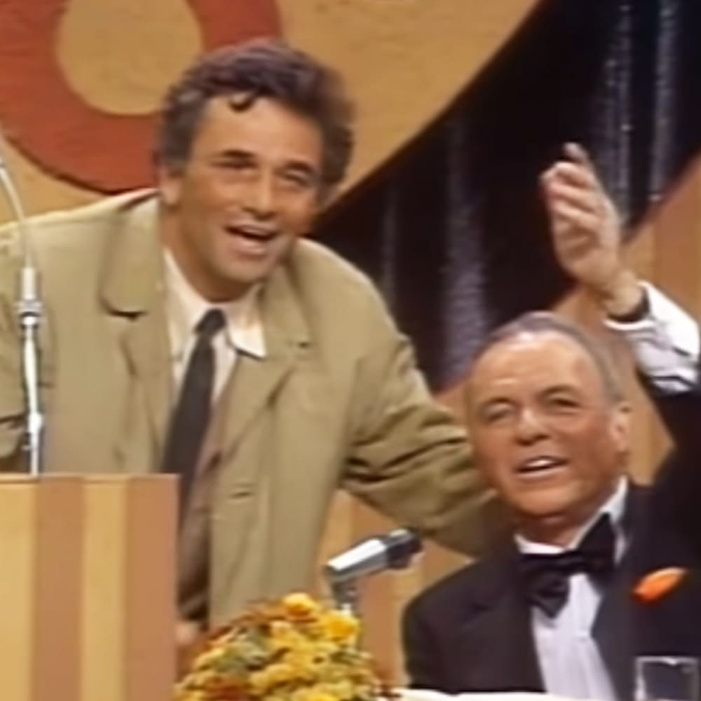 A middle-aged man wearing a beige blazer with dark tie stands beside an older adult male in a black tuxedo, both seated at a table with microphones and a glass on it. Behind them is a large abstract painting with warm tones and dark curtains.