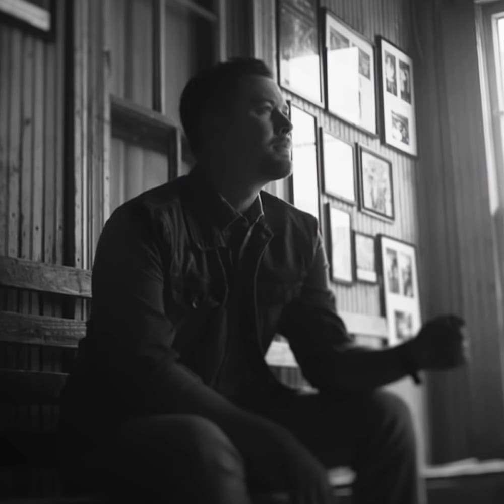An adult male seated on a wooden bench in an indoor setting with framed artwork, rustic wood paneling, natural light suggesting daytime.