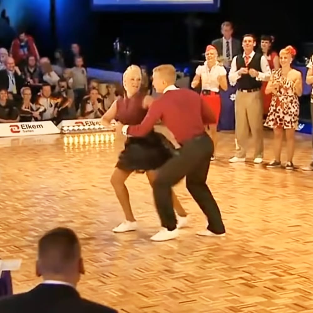 An indoor dance performance with a male adult in his 30s or 40s wearing a red long-sleeved shirt, dark pants, white socks, and white shoes. The setting is an artificial-lit venue with wooden flooring and spectators seated around the dance floor watching the performance.