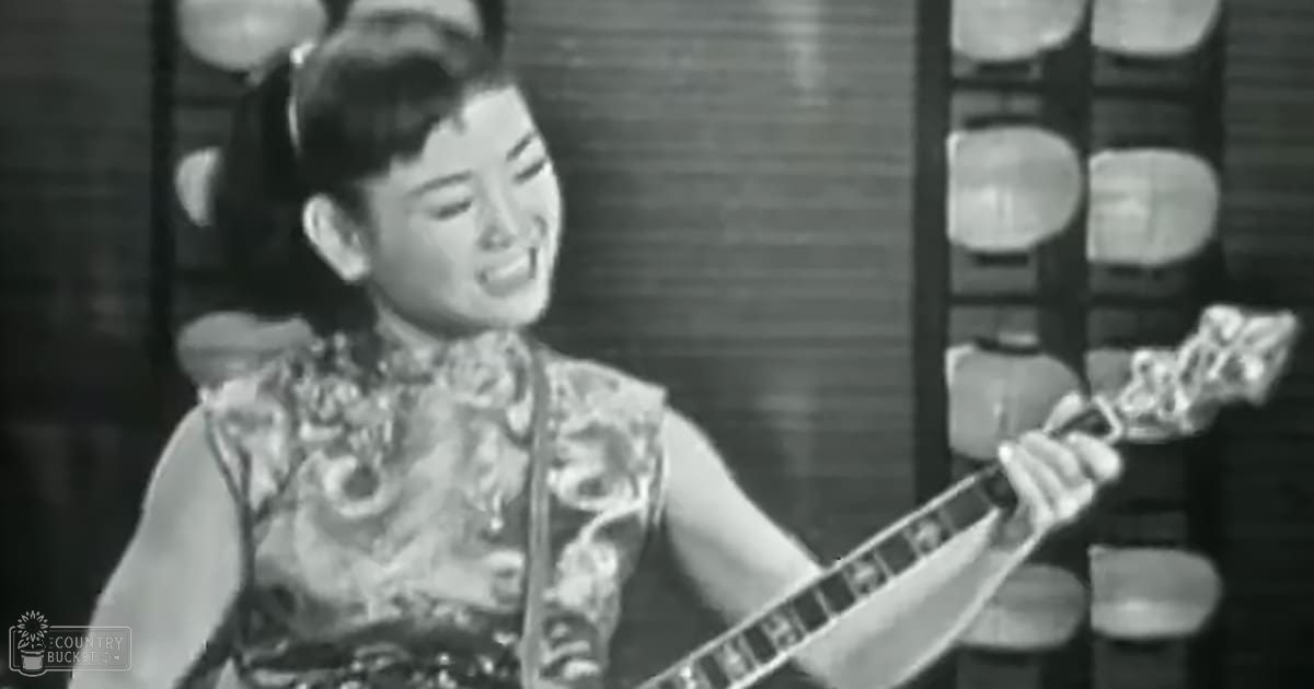 A young adult female in a traditional Chinese-style dress with intricate embroidery, holding a stringed instrument indoors during the day.