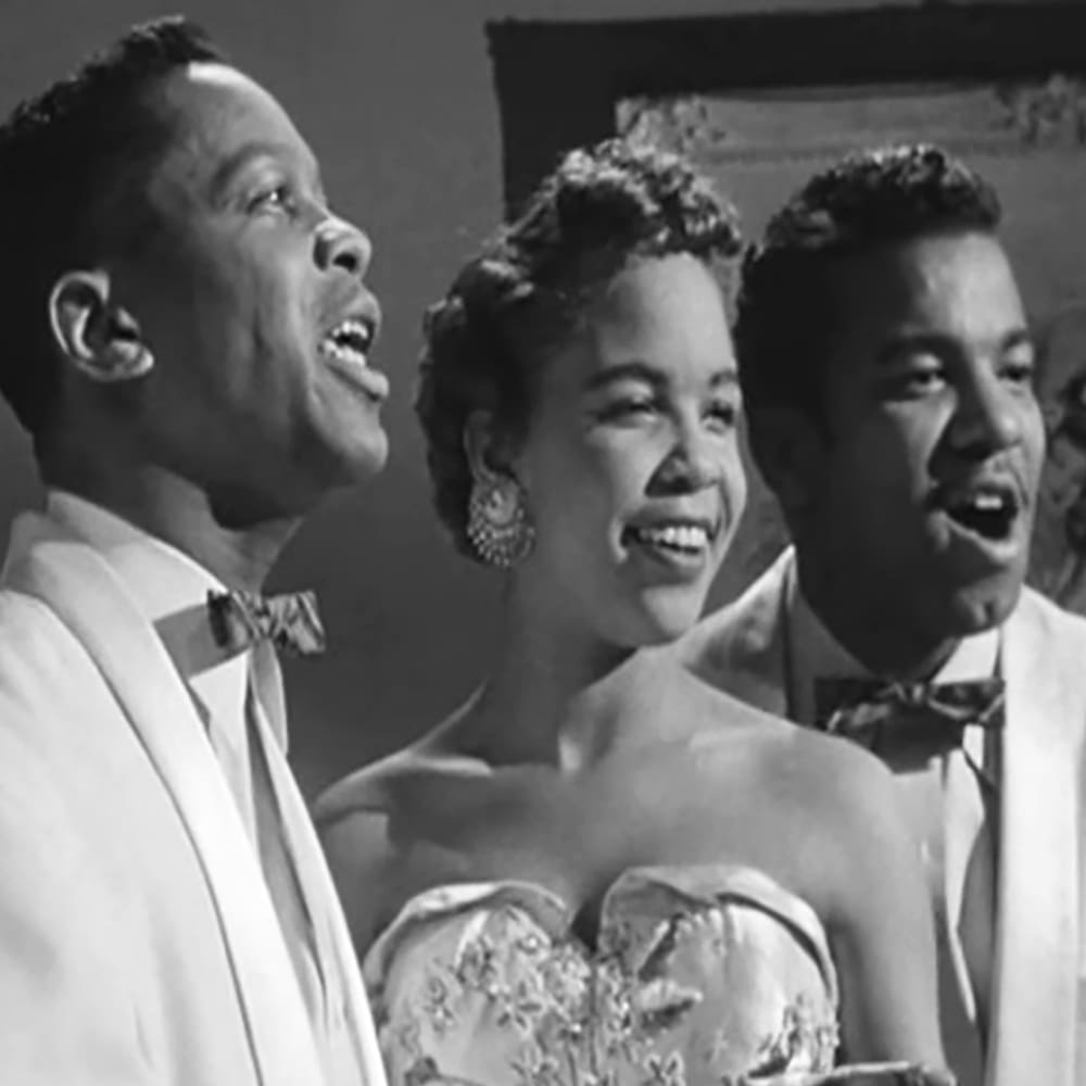 A mid-20th century America musical performance scene with a male in a white suit jacket and bow tie on the left, an adult female wearing an elegant floral dress in the center, and another male in similar attire on the right. The setting appears to be indoors at an event like a concert or performance venue.