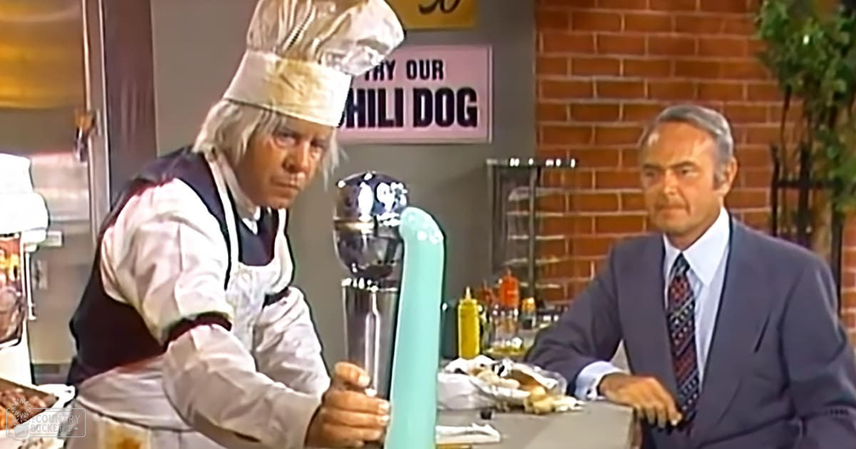 An adult male chef in a kitchen setting, wearing a white shirt under a dark vest and apron, holding a beer tap or dispenser. A seated older adult male in a light blue suit jacket is nearby.