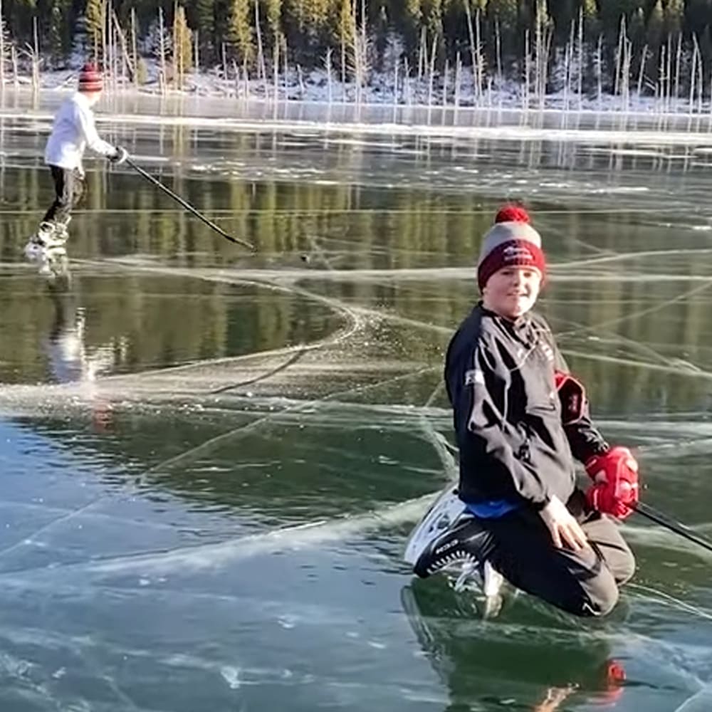 A young boy in a dark jacket and red beanie participates in ice fishing at a frozen lake surrounded by snow-covered trees.