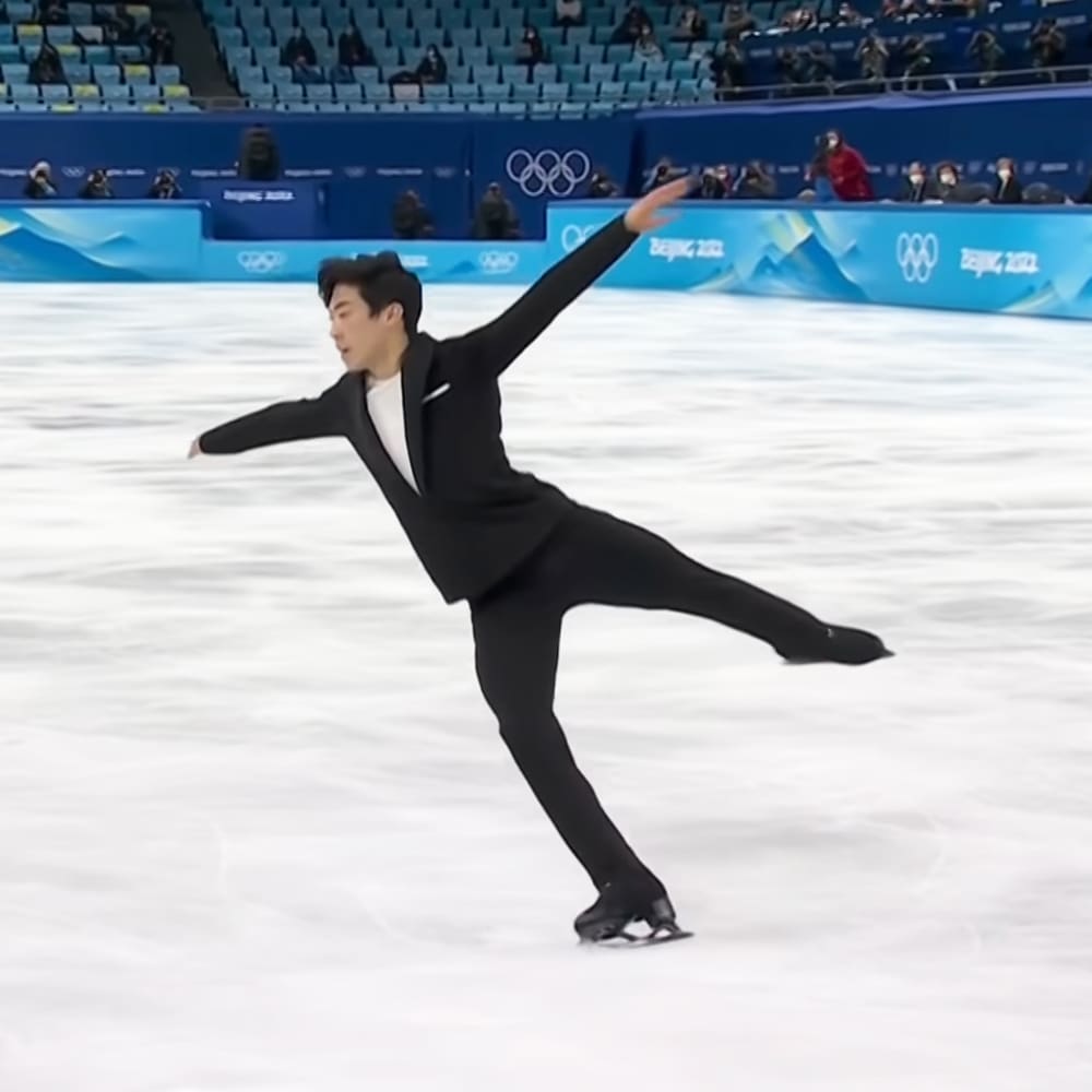 A young adult male figure skater in a black tuxedo with white shirt and black pants, performing at the Beijing 2022 Winter Olympics on an indoor ice rink.