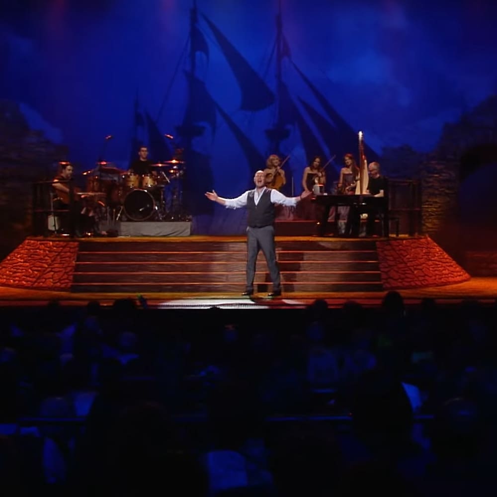 A male adult performer on a theater stage equipped with musical instruments, under theatrical lighting at night.