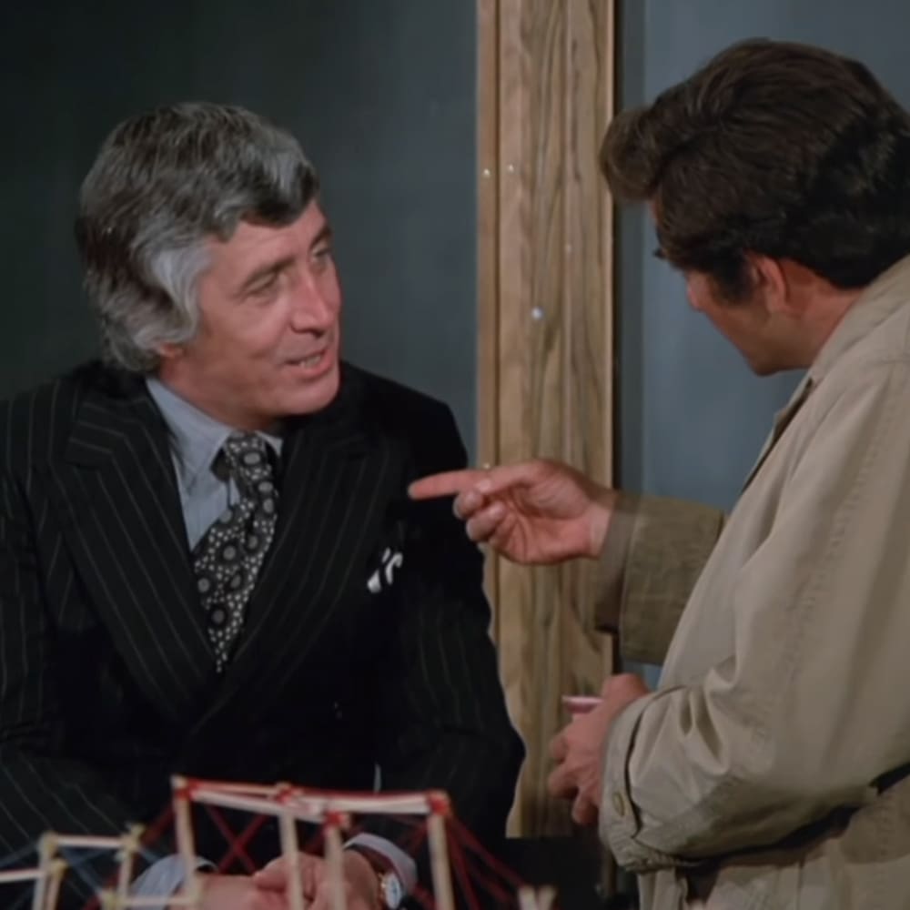 A middle-aged man wearing a dark pinstriped suit jacket, white shirt, and patterned tie sits next to a young adult male in a light-colored trench coat indoors, possibly in an office or meeting room setting.
