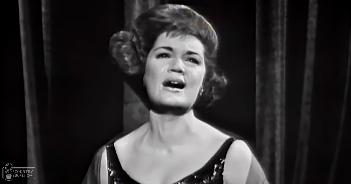 A middle-aged female singer wearing a dark mid-20th-century sleeveless garment with thin straps and low neckline, performing indoors under dim lighting.