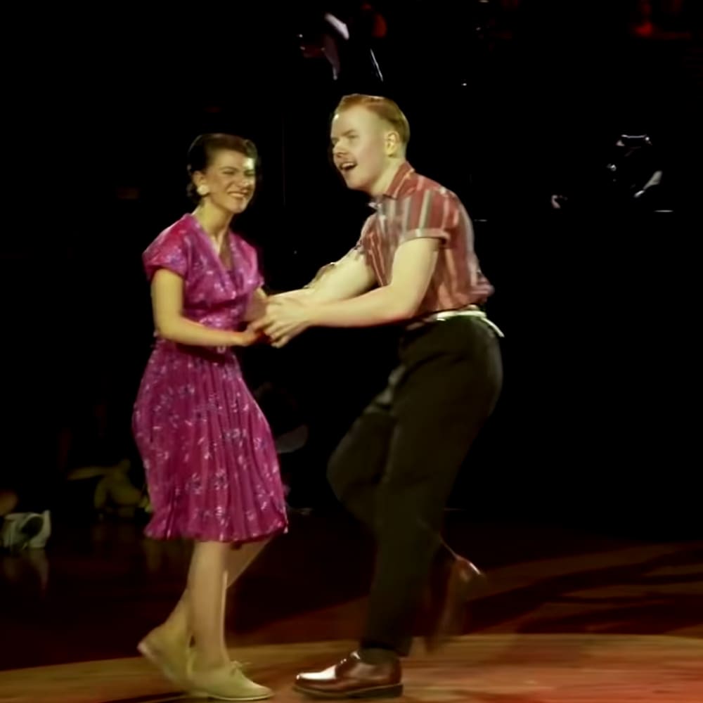 A young adult female dancer in a purple floral dress and ballet flats dances with a young adult male in a red-and-white striped shirt and dark green trousers on stage, under dim lighting suggesting a theatrical performance at night.
