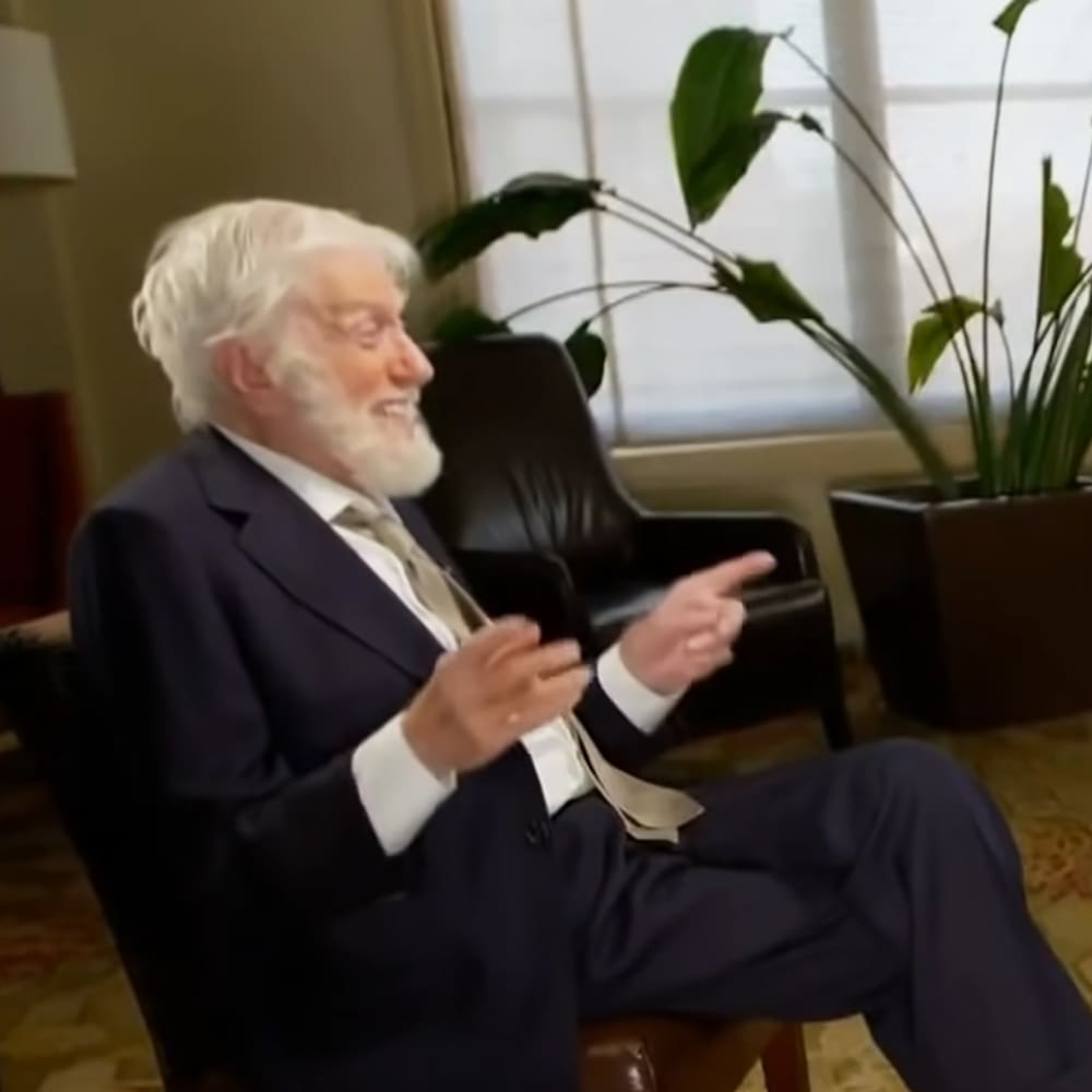 An older adult male in a dark blue suit jacket, white shirt, and light beige tie, seated in an office or formal interview room with black leather chairs and green potted plants.