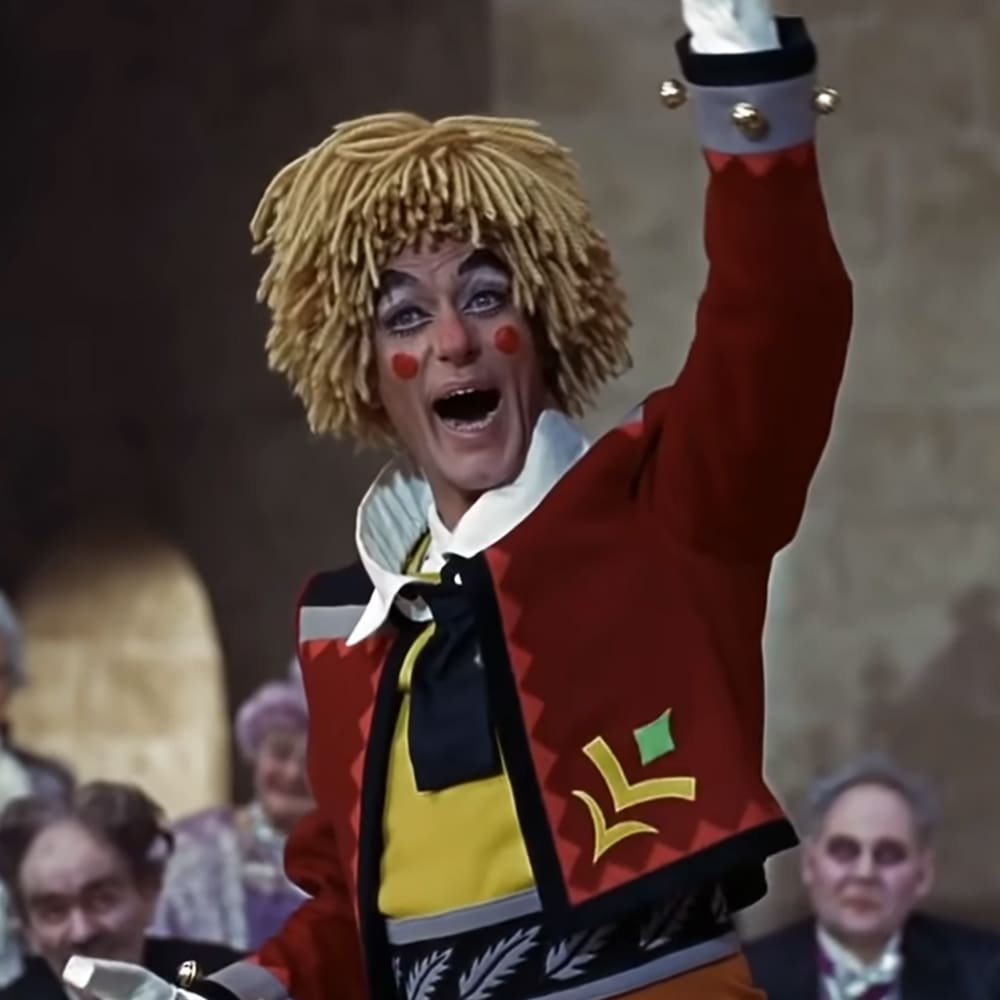 A male performer in a theatrical setting with an audience, dressed in a red jacket and yellow shirt, possibly inspired by circus performers or clowns; stone walls suggest a historical building.