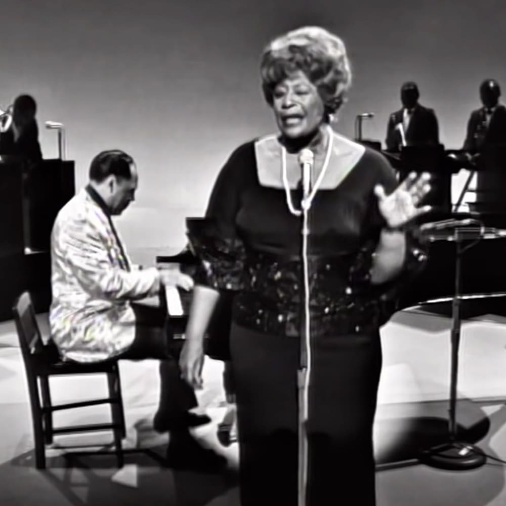 A woman singing in a studio setting with microphones and musical instruments, surrounded by adults in formal attire.