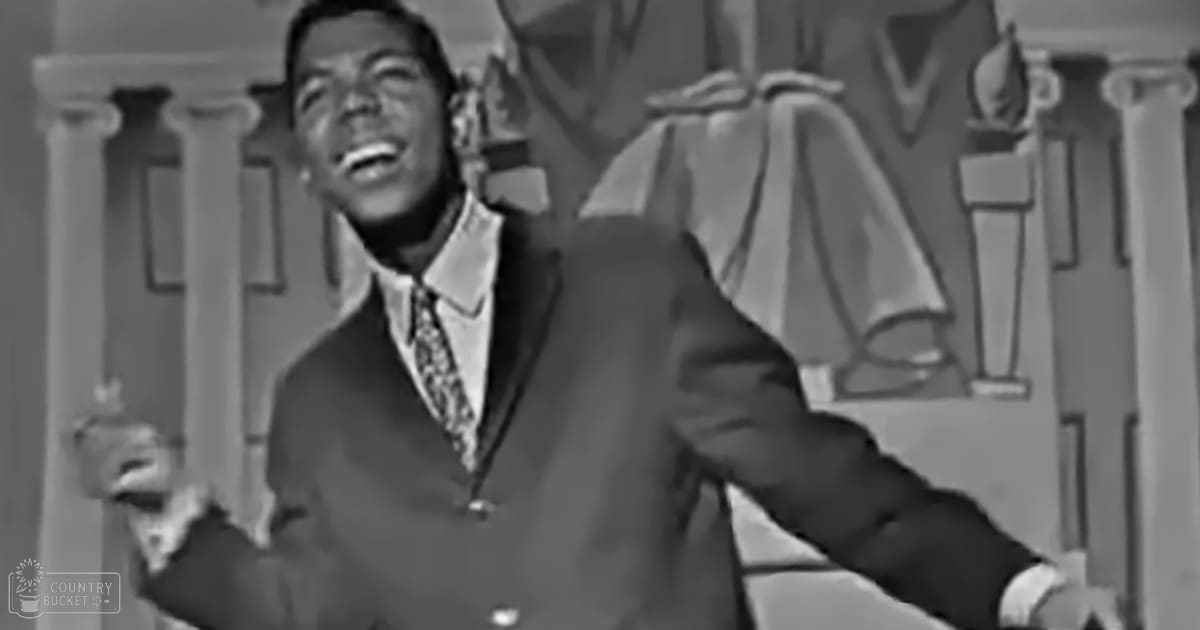 A young adult male in a dark double-breasted suit jacket with light-colored shirt and patterned tie stands against a decorative wall, possibly in a performance space.