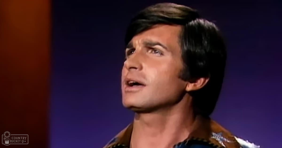 A young adult male wearing a patterned blue and brown collared shirt with short sleeves, standing against a dark blue-purple gradient background in an indoor setting.