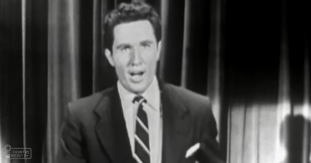 A young adult male in a dark suit jacket, white dress shirt, and striped tie stands indoors with vertical blinds or curtains in the background.