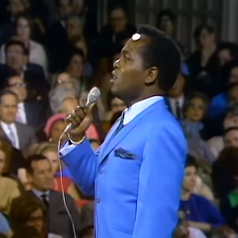 A middle-aged adult male singer in a bright blue suit jacket and tie performs into a microphone at an indoor event with seated audience members dressed in formal attire.