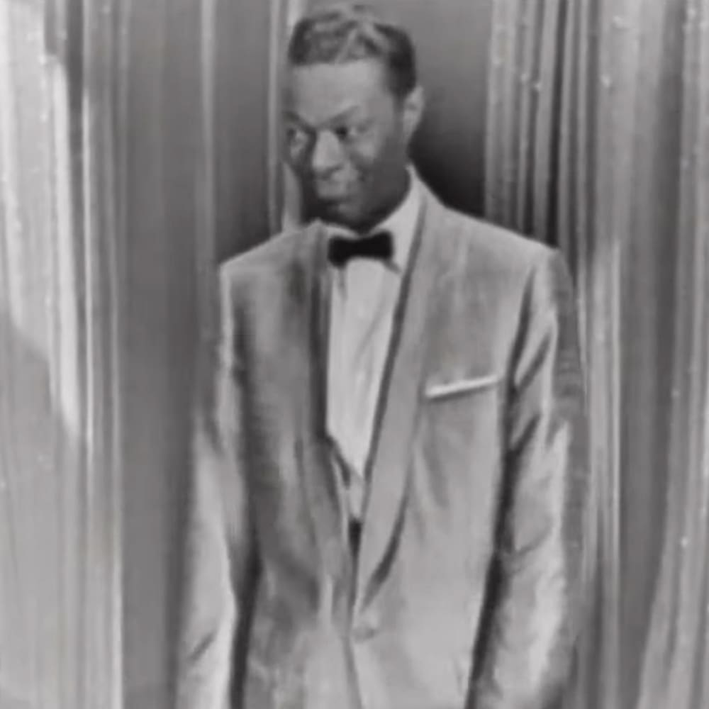 An adult male in a light-colored suit jacket with a white shirt, black bow tie, and pocket square or small accessory, standing indoors against vertical wooden panels.