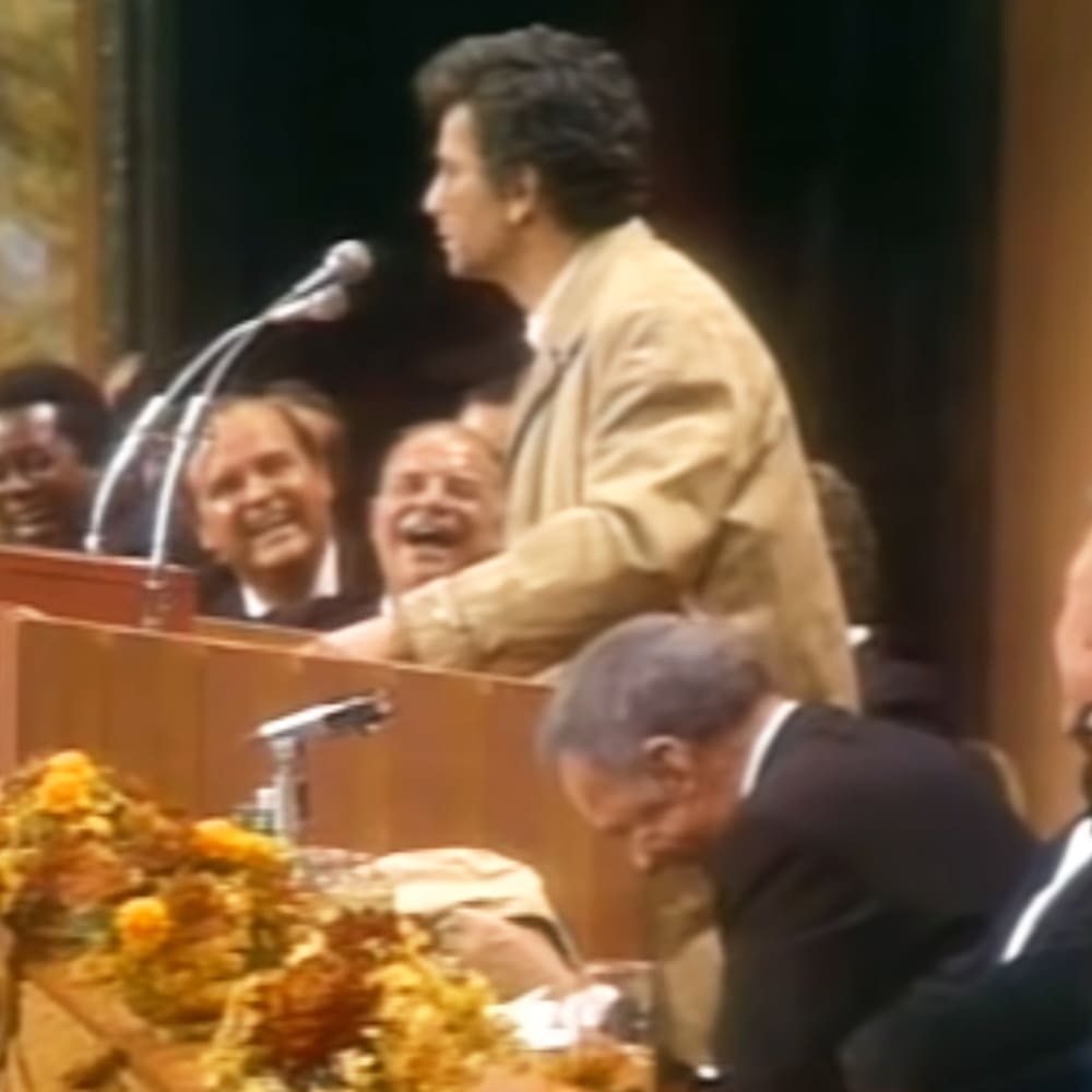 An adult male speaker wearing a light brown jacket and white shirt at an indoor podium event, with an audience in formal attire; dark curtains and stage setup suggest a conference or gathering setting.