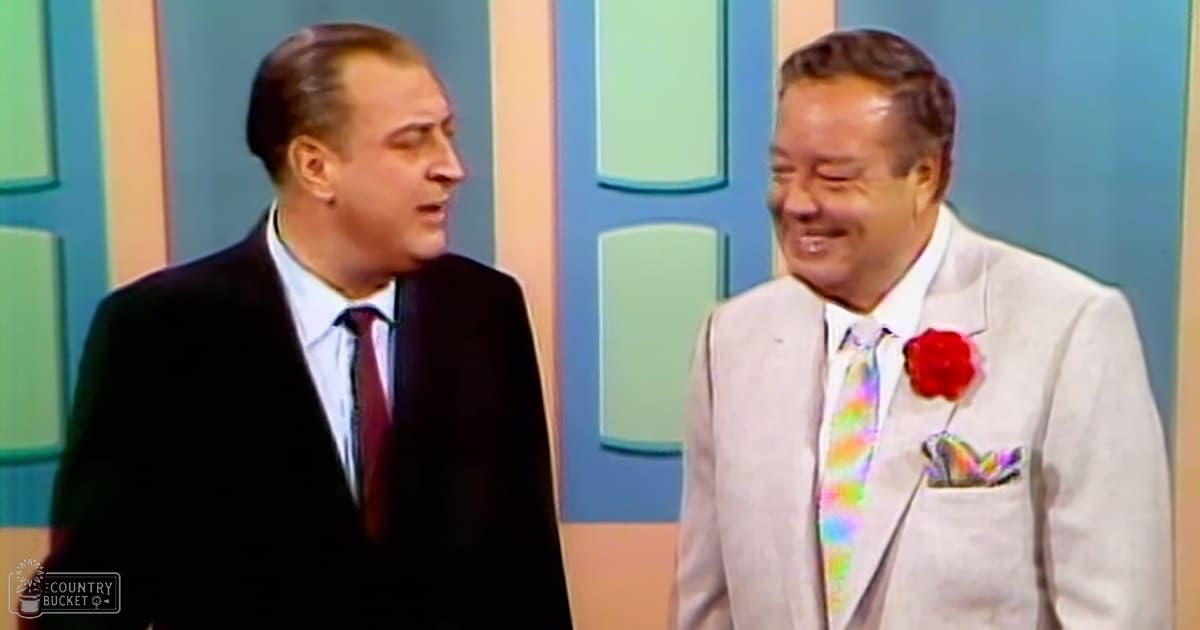 Two men in a studio setting wearing formal suits with colorful ties; one has a red tie, the other wears a rainbow patterned tie and a red rose lapel pin.