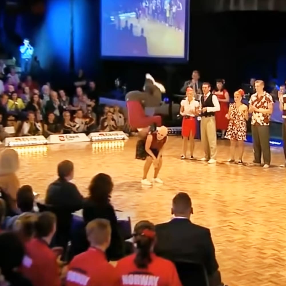 A young adult female performer in a sleeveless maroon top and red shorts or skirt, performing a dance move at an indoor venue with seated audience members wearing casual attire.