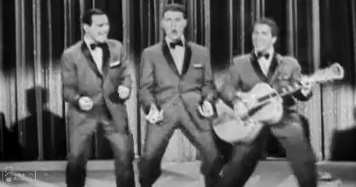 Adult males dressed formally in dark suits with white shirts and black bow ties on a stage with vertical curtains, suggesting an indoor venue from the mid-20th century.
