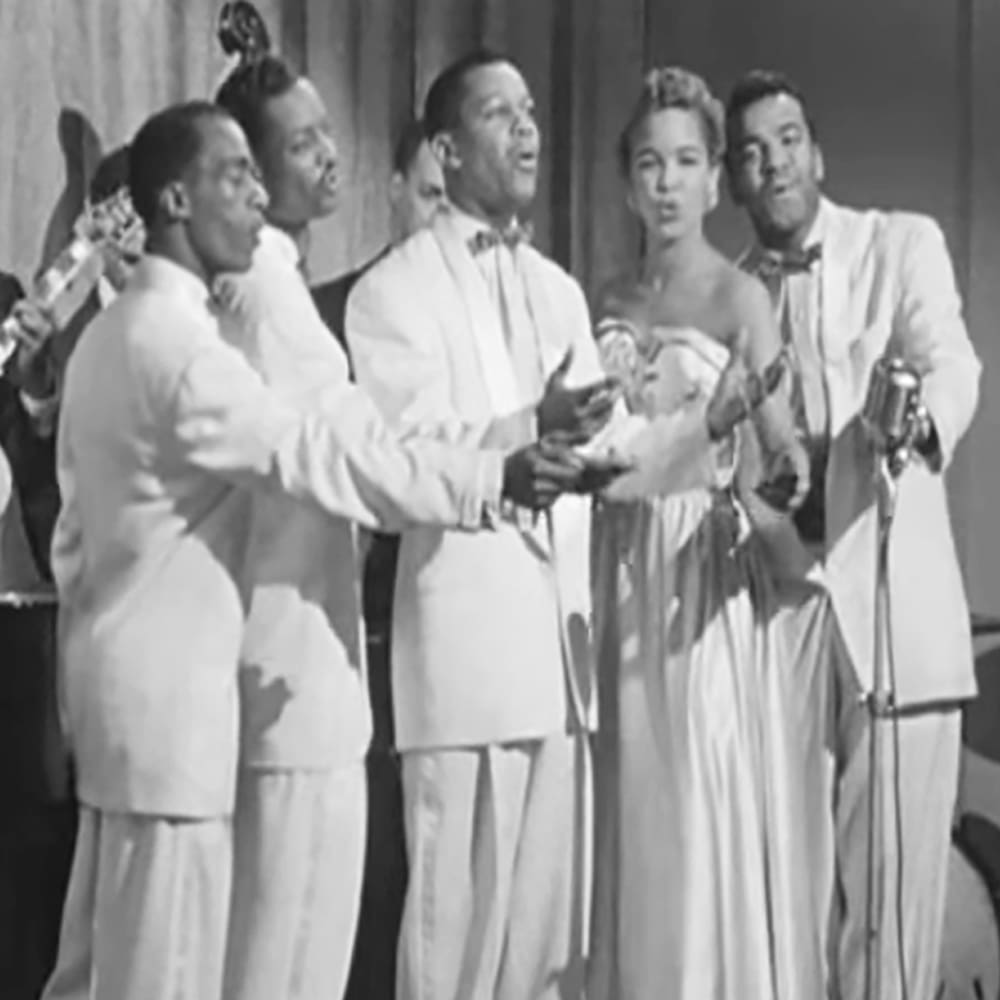 A mid-20th century musical performance group in a formal indoor setting with microphones on stands.