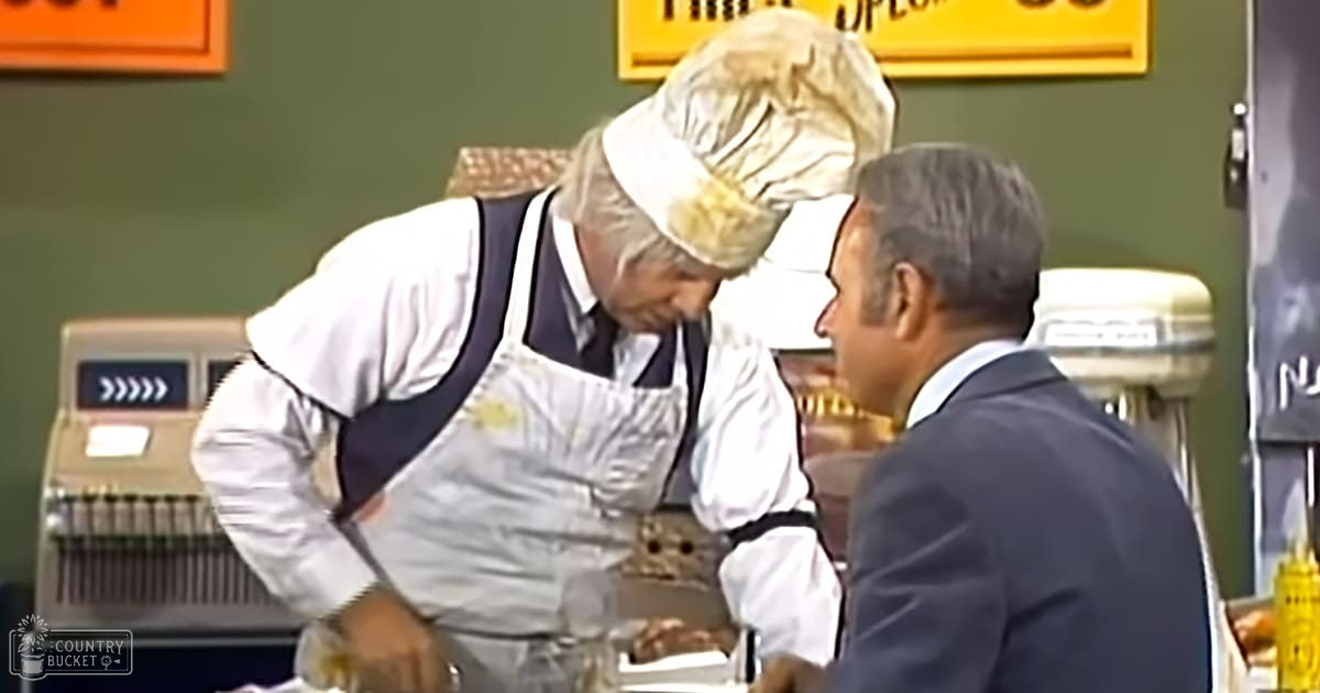 A middle-aged male in a white shirt with rolled-up sleeves, dark blue overalls, and gray hair is interacting at what appears to be a bakery or deli counter. Next to him stands an adult male in a dark suit jacket and light-colored collared shirt.