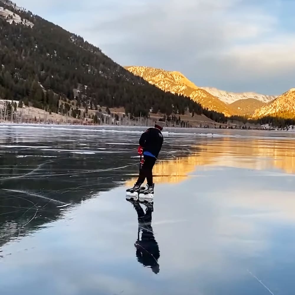 An adult male ice skating on a frozen lake surrounded by mountains, wearing a black jacket with red accents and white ice skates in winter light.