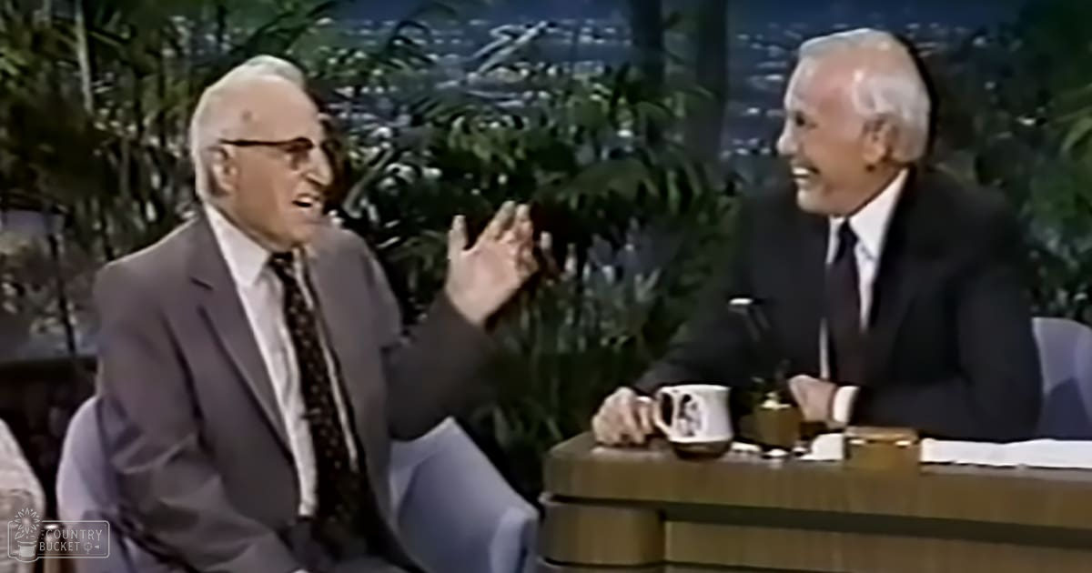Two older adult men conversing in a talk show studio with mid-20th century style attire, greenery and city lights in the background.