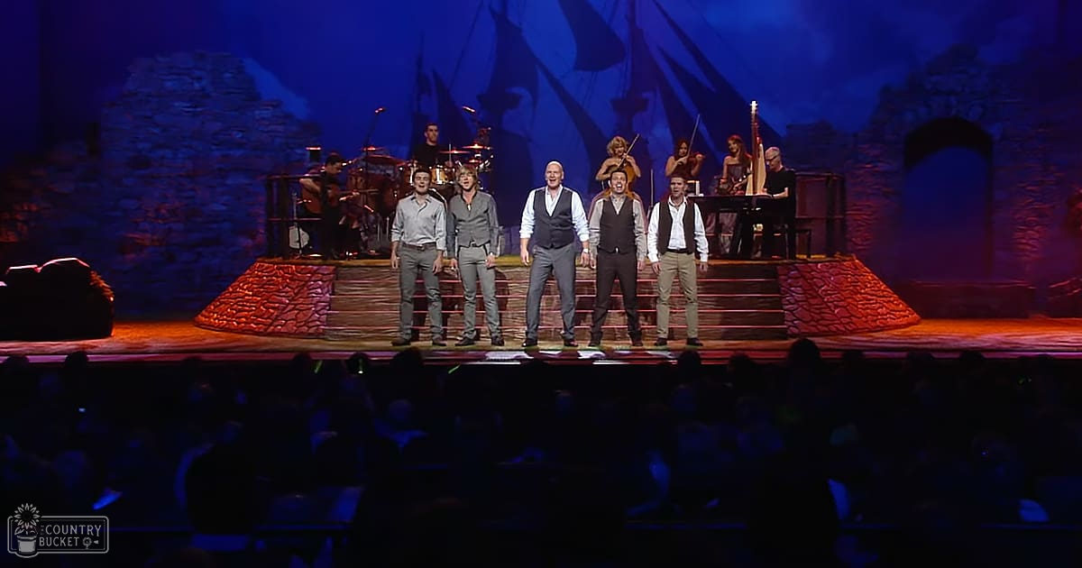 An indoor theatrical performance with dimly lit historical or fantasy-themed stage set against an ancient stone structure backdrop, with various musical instruments on a red carpeted floor and performers in period-appropriate attire.