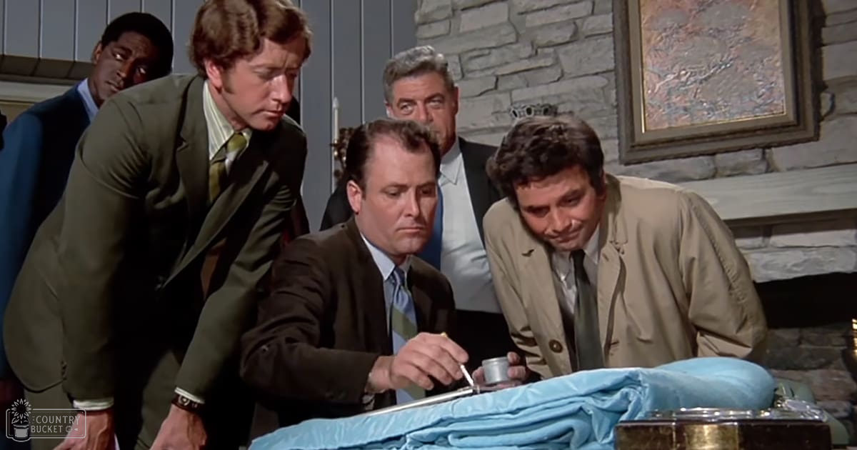 An adult male in a dark brown suit jacket and light blue striped tie examines an object under bright, even lighting in what appears to be an office or study room with a stone wall backdrop and framed artwork.