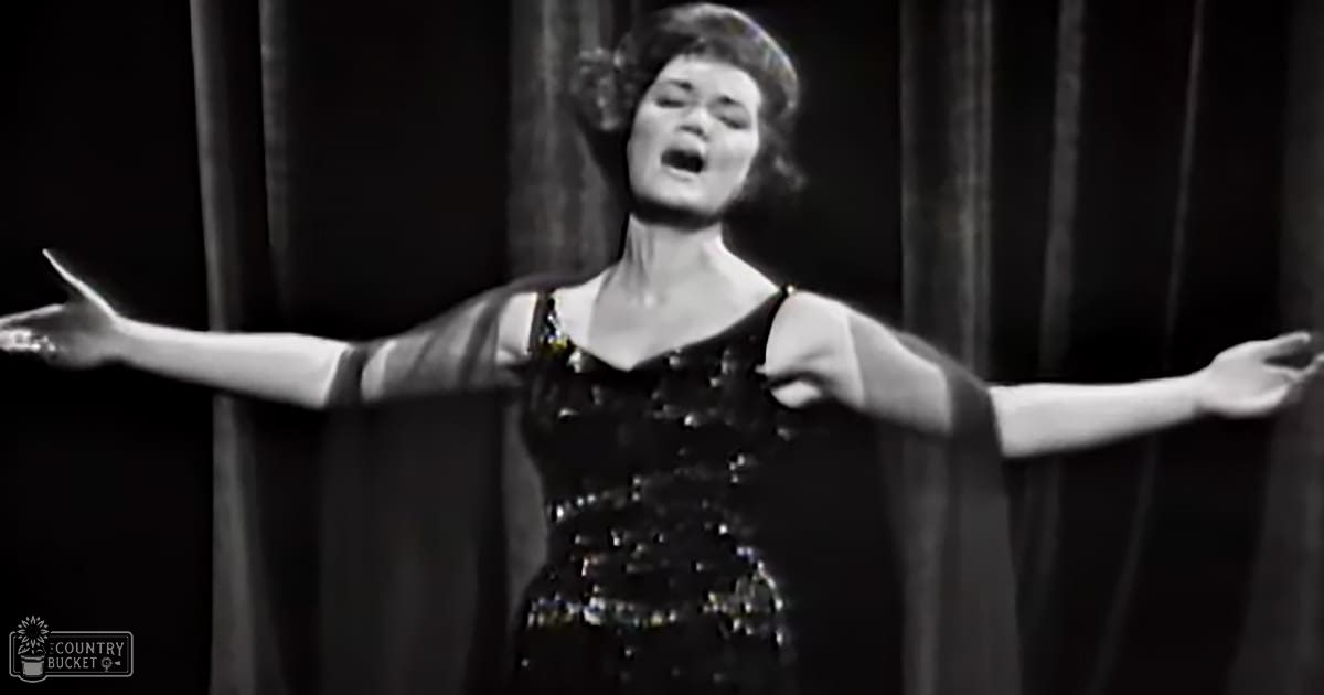 Adult female performer wearing a form-fitting, sequined black dress with wide puffed sleeves, set against dark curtains indoors.