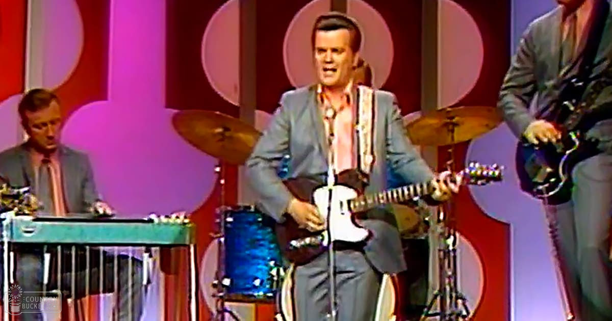 An adult male performer in a light gray suit and orange patterned shirt, accompanied by two other adult males dressed similarly on stage with musical instruments, under vibrant red and purple lighting.