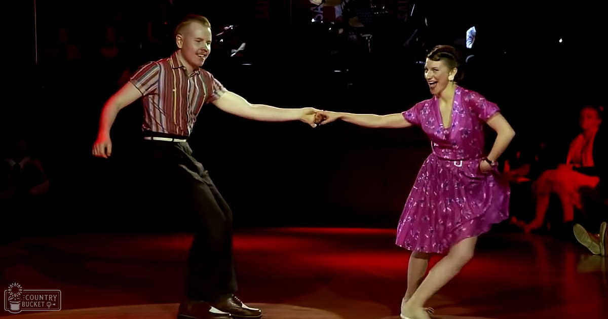 A dance performance scene indoors with stage lighting, possibly a studio or venue at night; young adult male in striped shirt and trousers, young female in shiny purple dress.