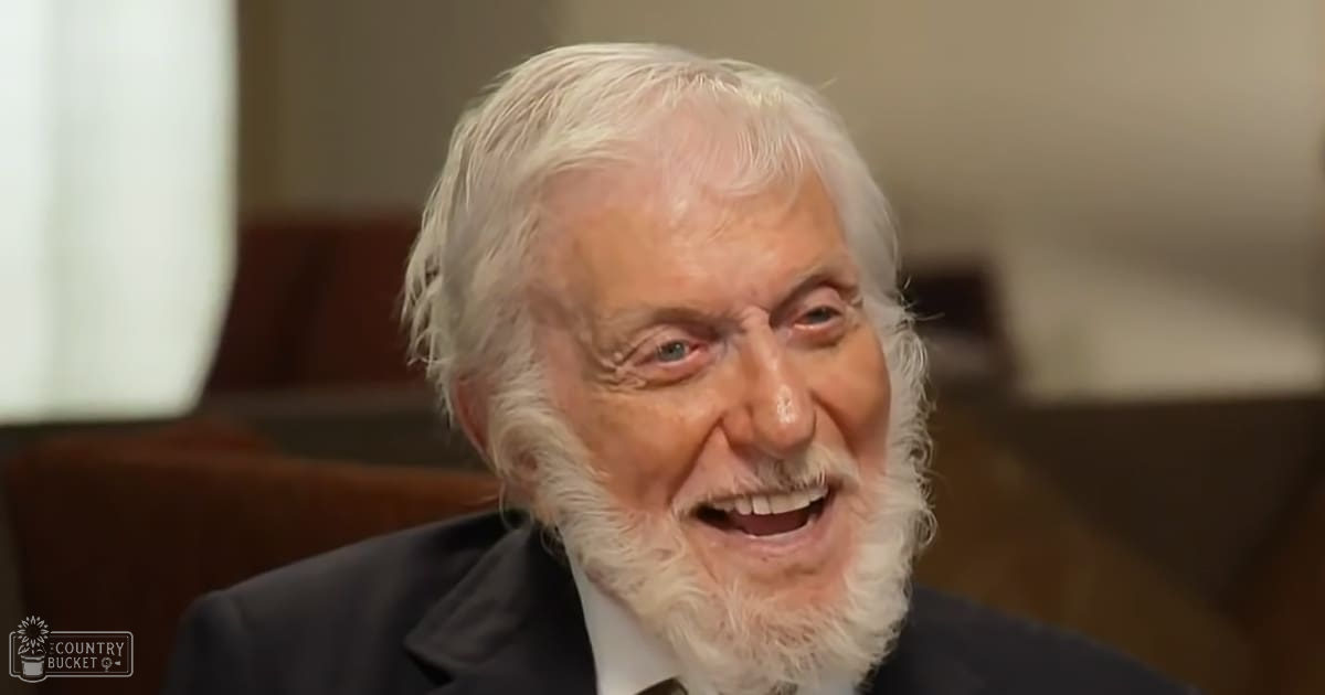 An older adult male in a formal setting, wearing a dark-colored suit jacket with a white shirt and a tie made of silk or similar material from the mid-20th century. Indoors, possibly an office or formal room, natural light suggesting daytime.