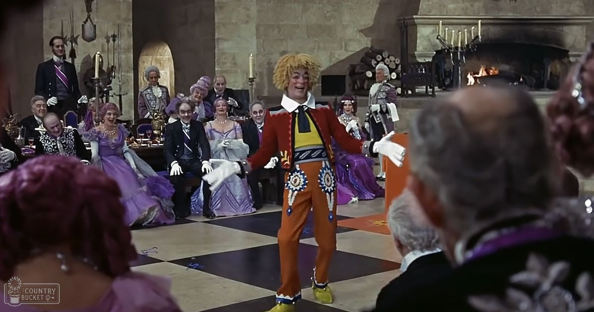 A performer in a vibrant red jacket with gold and blue accents stands center stage against an opulent indoor setting resembling a grand ballroom or castle hall, surrounded by audience members dressed in formal historical attire.