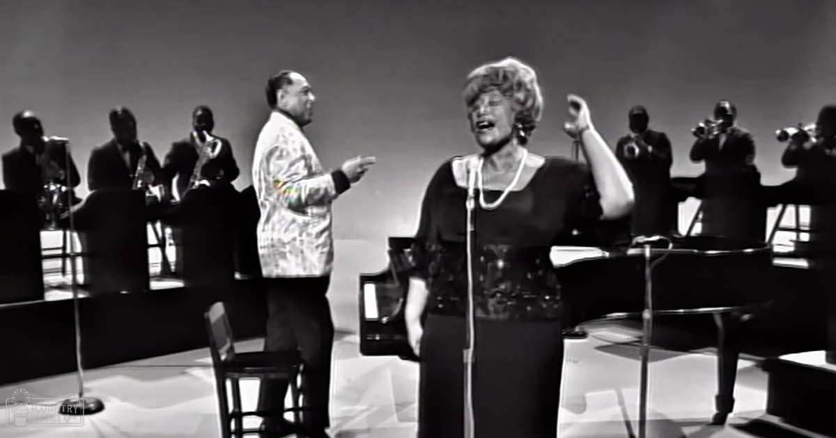 A woman in a patterned black dress sings on stage, accompanied by a conductor wearing a light-colored paisley jacket and dark pants; setting suggests an indoor performance venue with simple backdrop.