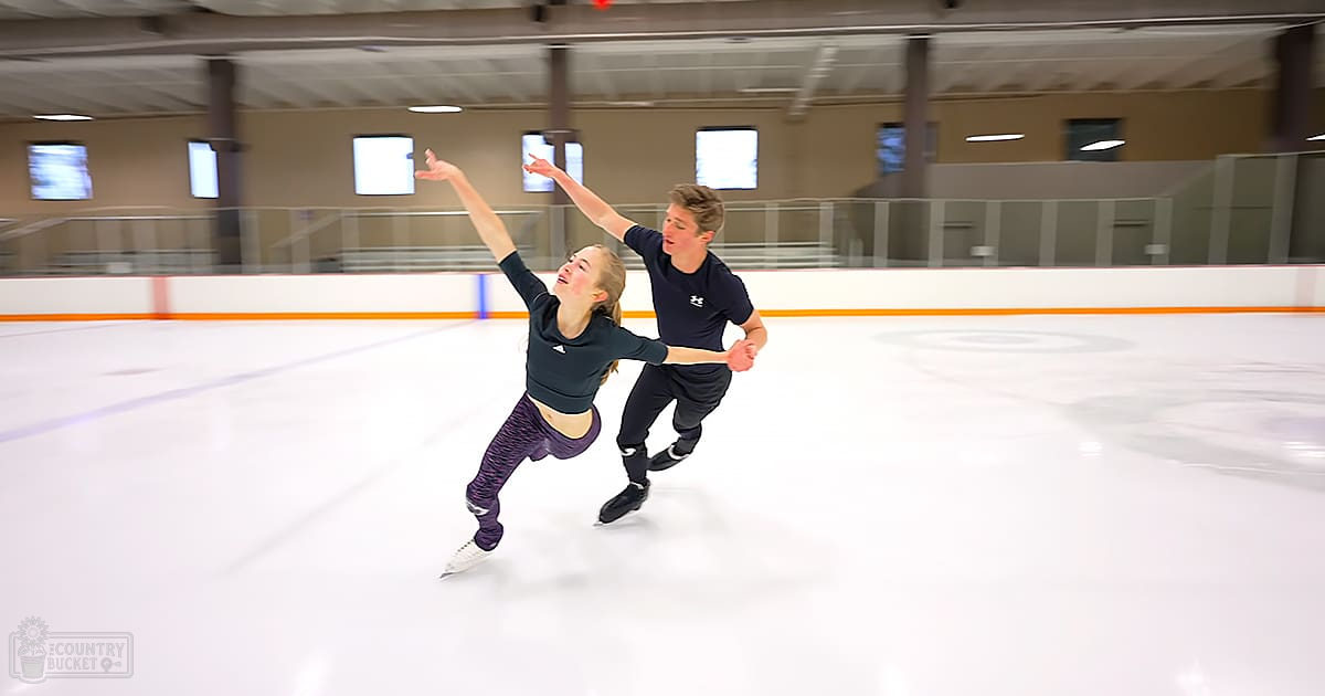 A young couple ice skating indoors at an indoor rink with white flooring and orange boundary lines, natural light from windows indicating daytime.