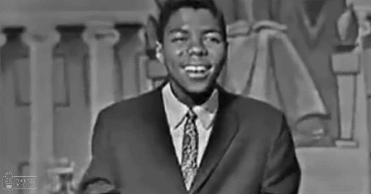 A young adult male wearing a tailored dark suit, white shirt, and patterned tie stands indoors against a backdrop with columns and part of the letter 'M'. The setting is well-lit with studio lighting.