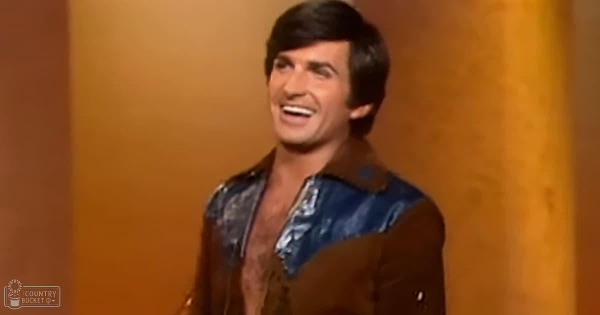 Young adult male in a brown jacket with blue patterned shirt, indoors with warm-toned walls and artificial lighting.
