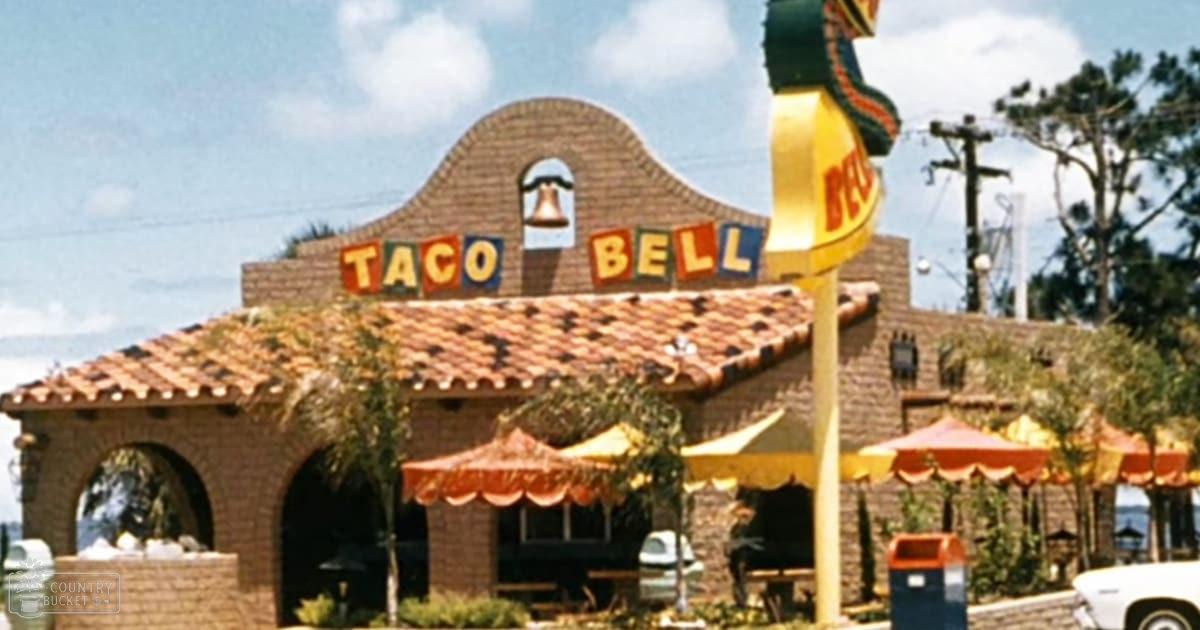 An outdoor view of a Taco Bell restaurant with colorful umbrellas, brick building, parked cars, clear sky, and trees in the background on a sunny day.