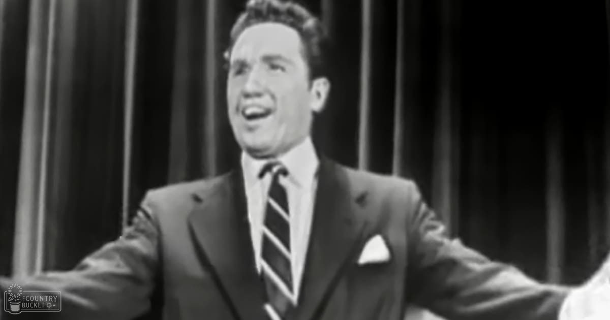 A young adult male in a formal suit with a striped tie, standing against vertical blinds or curtains on an indoor stage or studio backdrop.