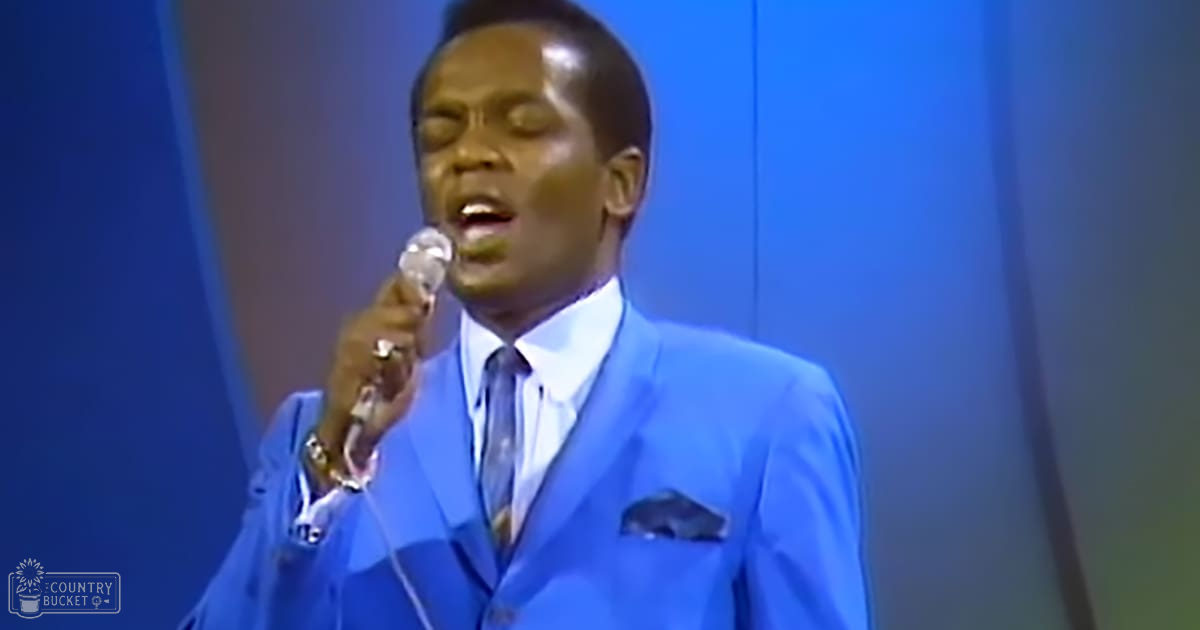 A middle-aged male singer wearing a bright blue suit jacket with pocket square, white dress shirt, and dark tie, singing into a microphone in a studio or stage setting.