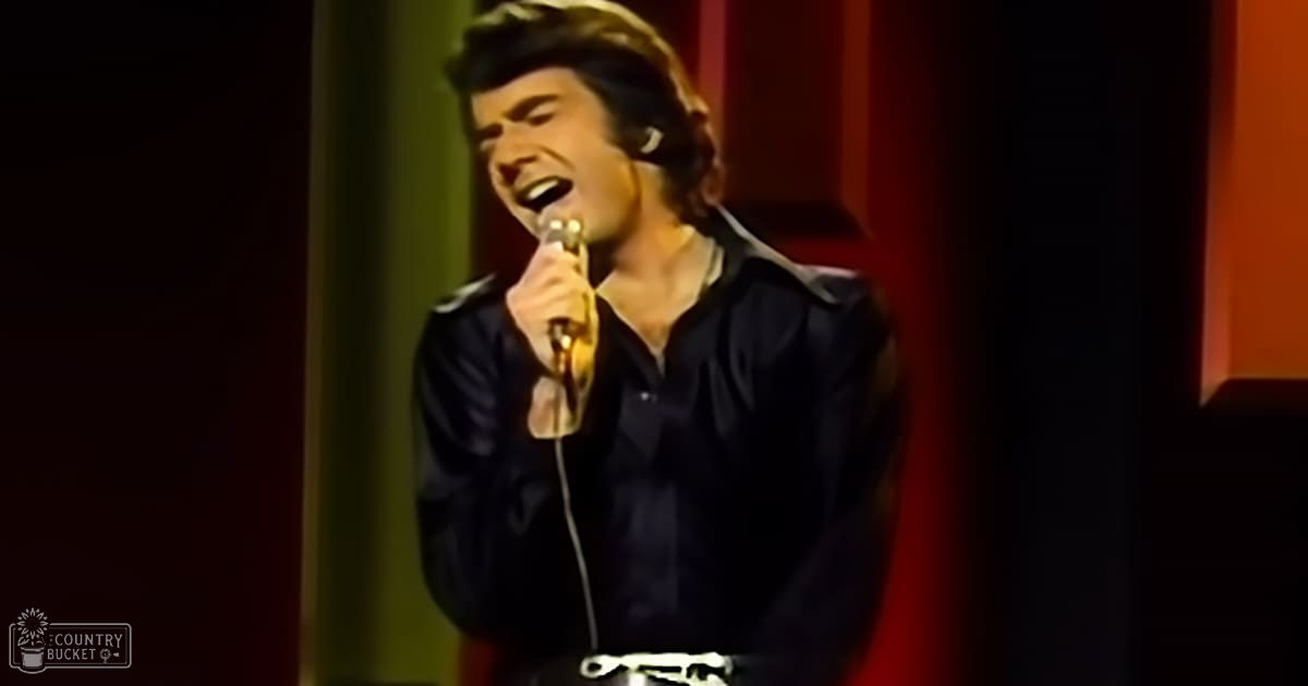 An adult male performer singing into a microphone, wearing a dark-colored 1970s-style button-up shirt on stage with red and green vertical panels in the background.