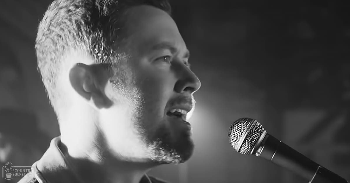 A young adult male singer wearing a light-colored jacket performs into a microphone against a dark backdrop with dramatic lighting in an indoor setting.