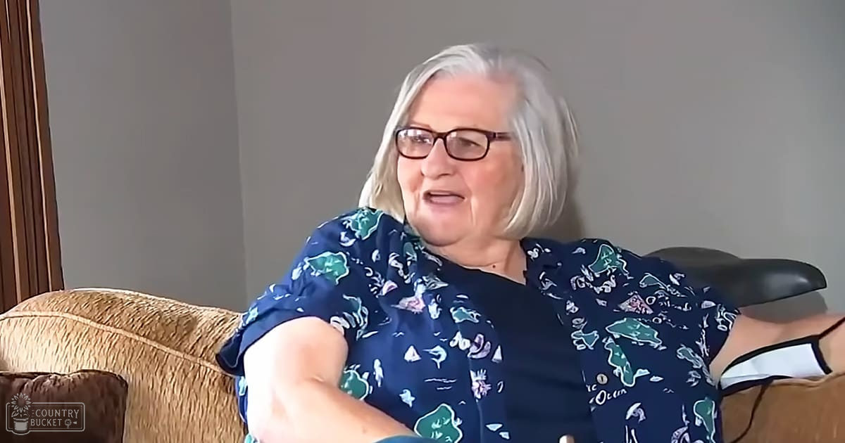 An older adult female seated indoors wearing a blue floral-patterned blouse with beige couch in the background, no natural light visible.